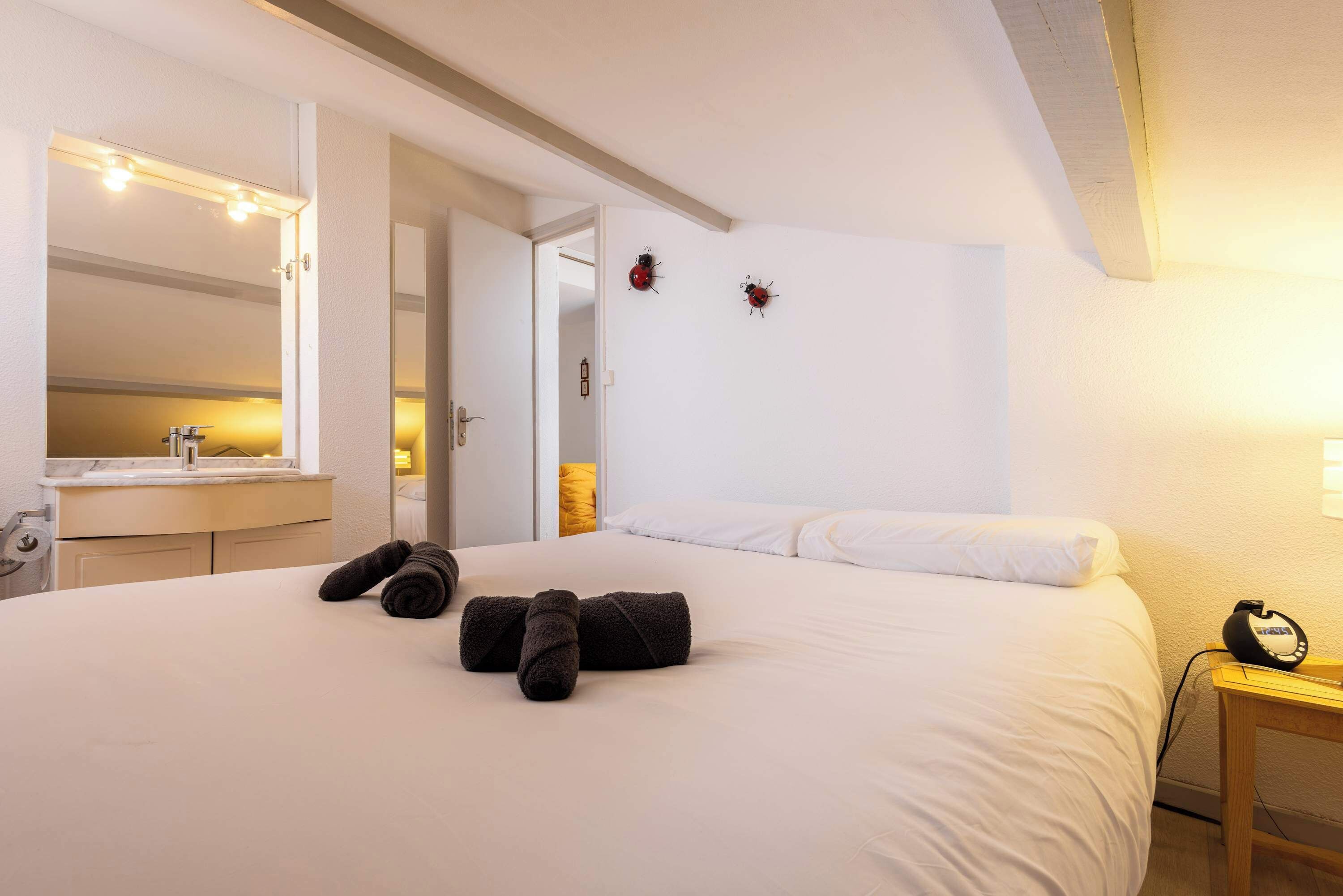 Habitación de hotel con cama, mesita de noche con lámpara, espejo con luces y puerta que da a otra habitación.