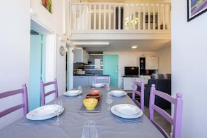 A dining area with a table set for four, chairs, a kitchen in the background, and a balcony with white railings.