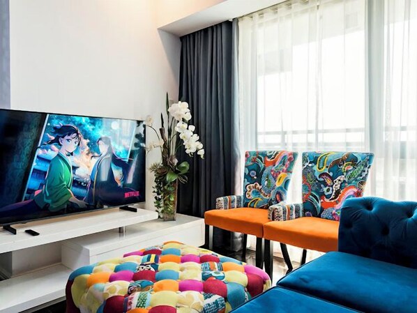 A modern living room with a flat-screen TV, colorful chairs, a blue sofa, and a vibrant ottoman.
