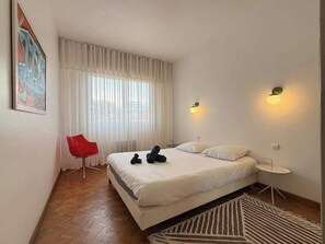 Un dormitorio con cama, una silla roja, una mesita y un cuadro en la pared.