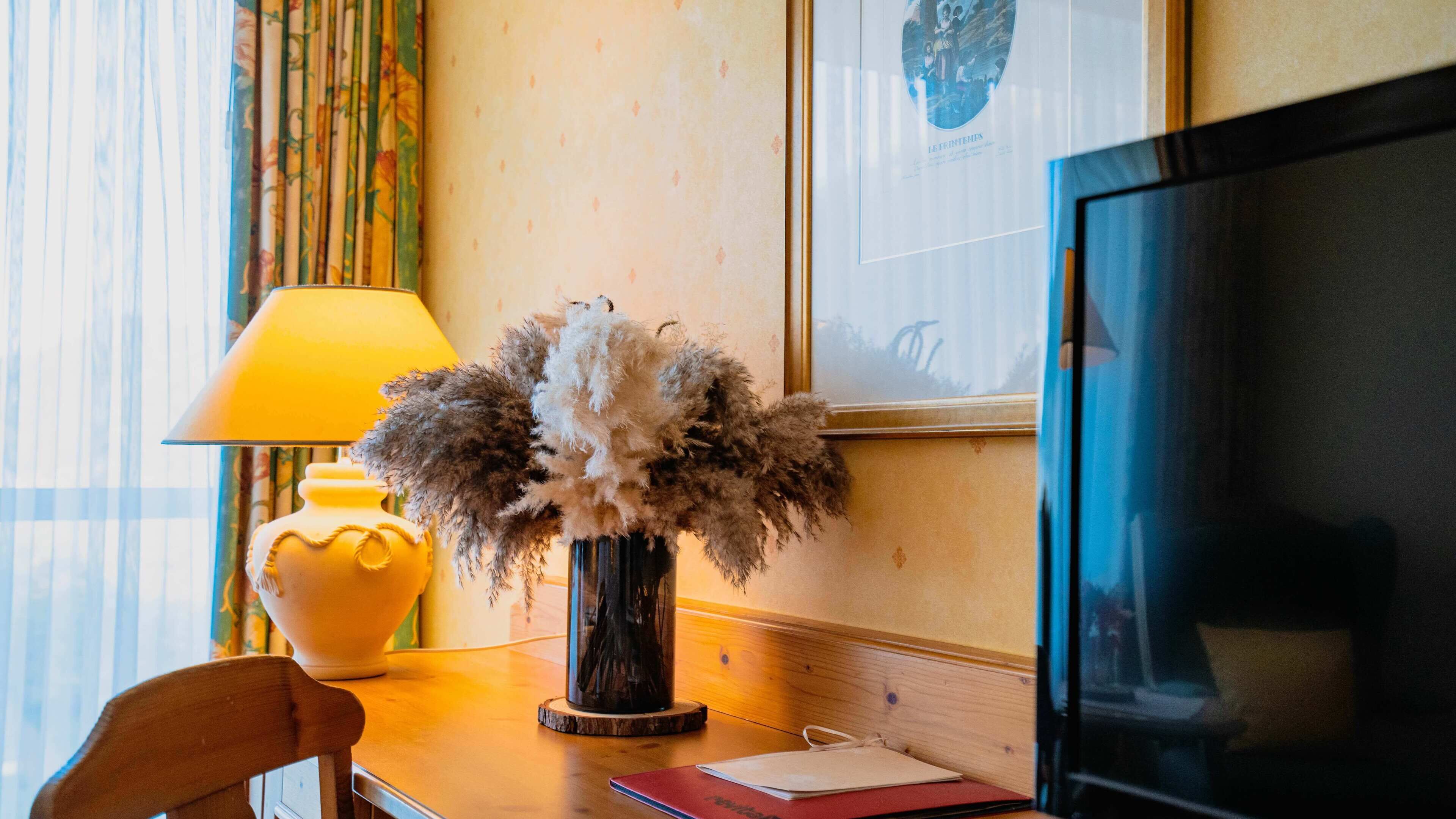 Ein Hotelzimmer mit Holzschreibtisch, einer Vase mit Trockenblumen, einer gelben Lampe, einem gerahmten Bild an der Wand und einem Fernseher.