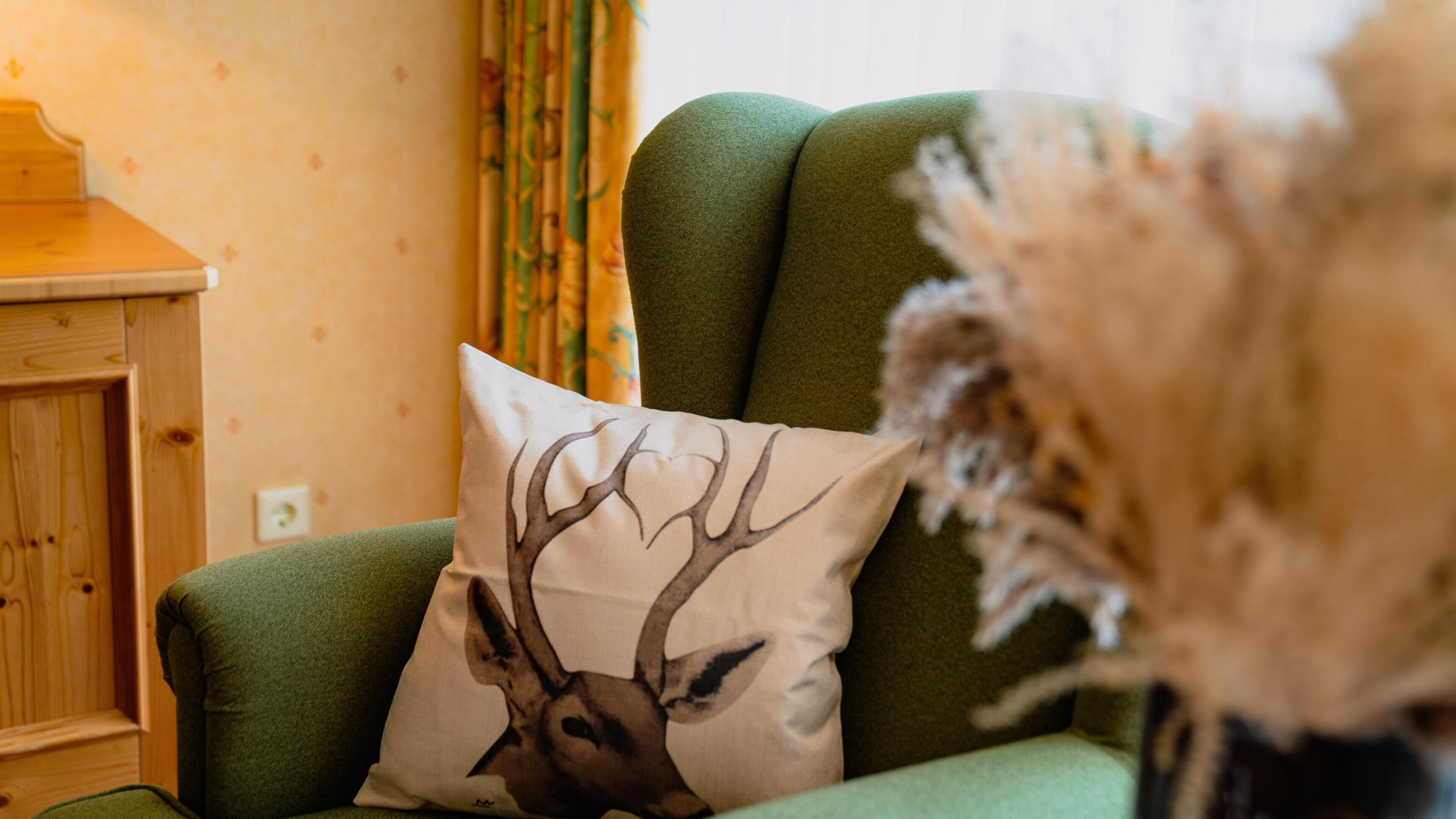 A green sofa with a deer print pillow, a wooden cabinet, and a vase with dried flowers.