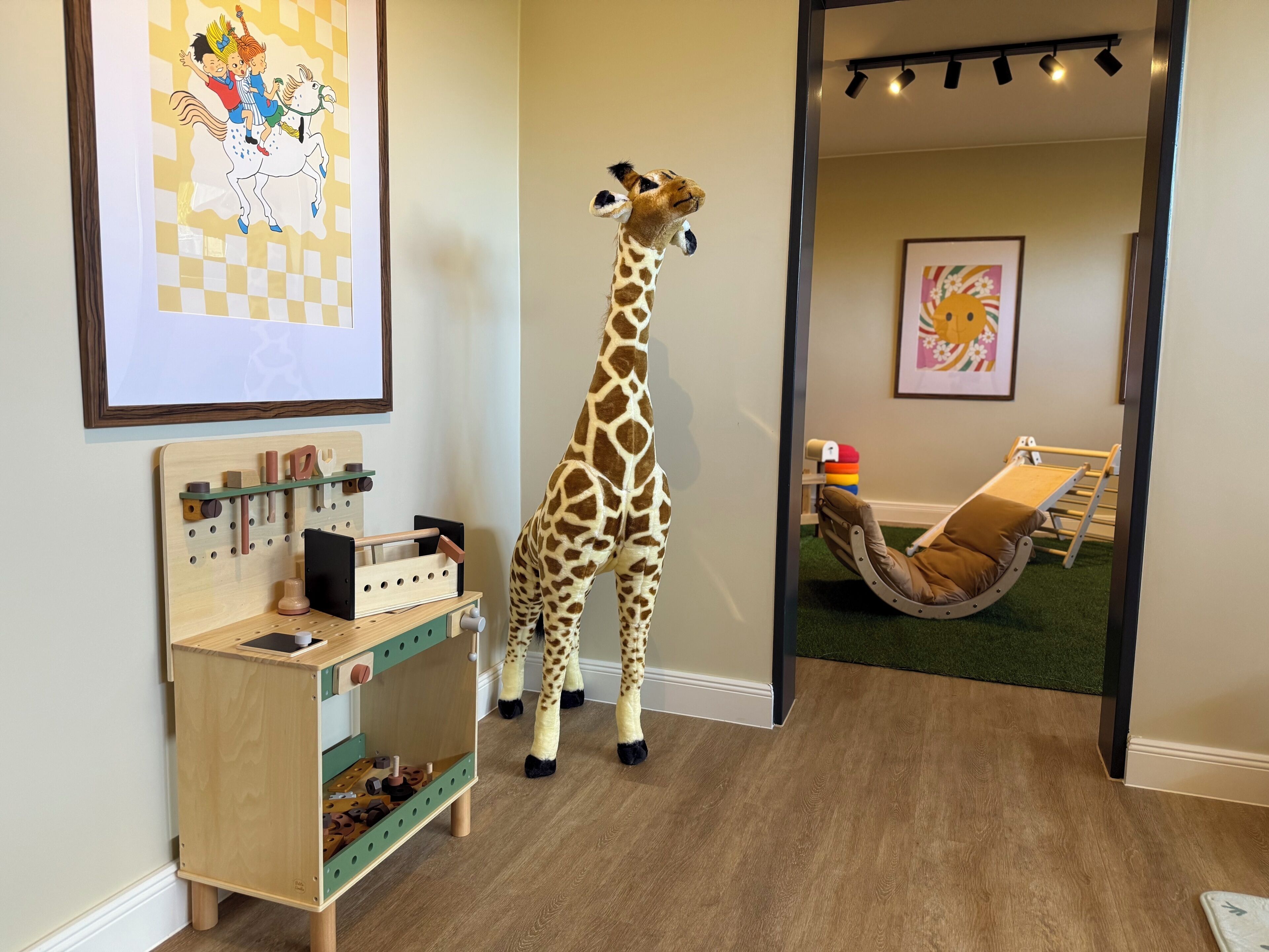 Une statue de girafe dans une pièce où se trouvent un parc pour enfants, un meuble de rangement pour jouets fixé au mur et un hamac.