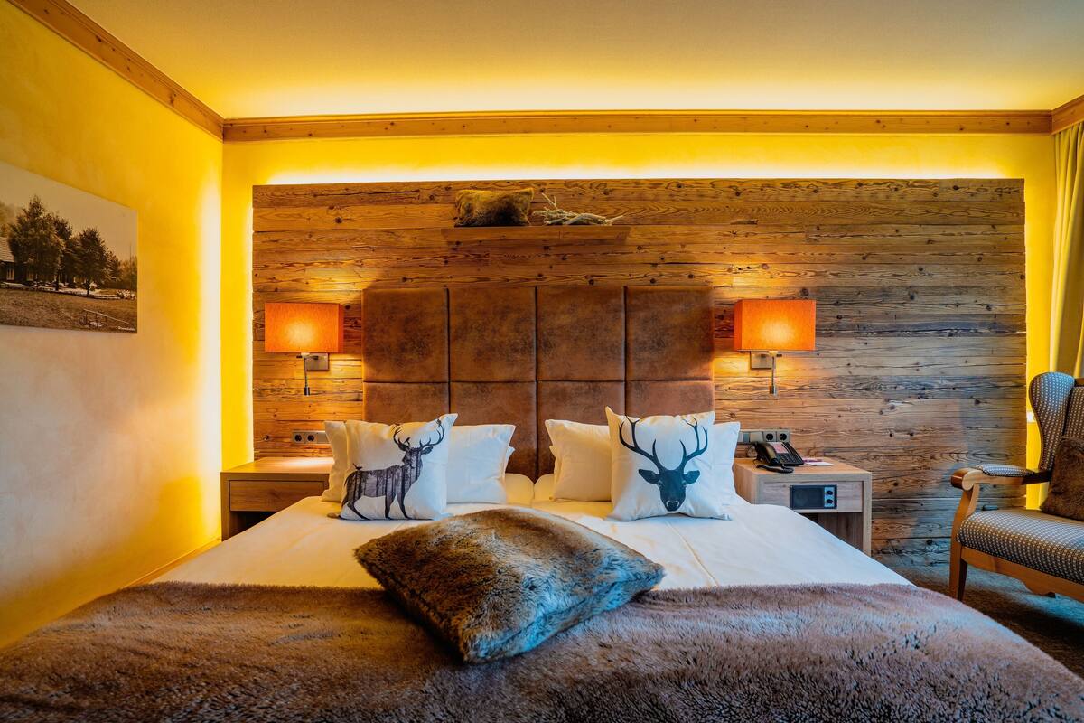 Una habitación con cabecera de madera, cama con ropa blanca, dos lámparas de noche y una silla.