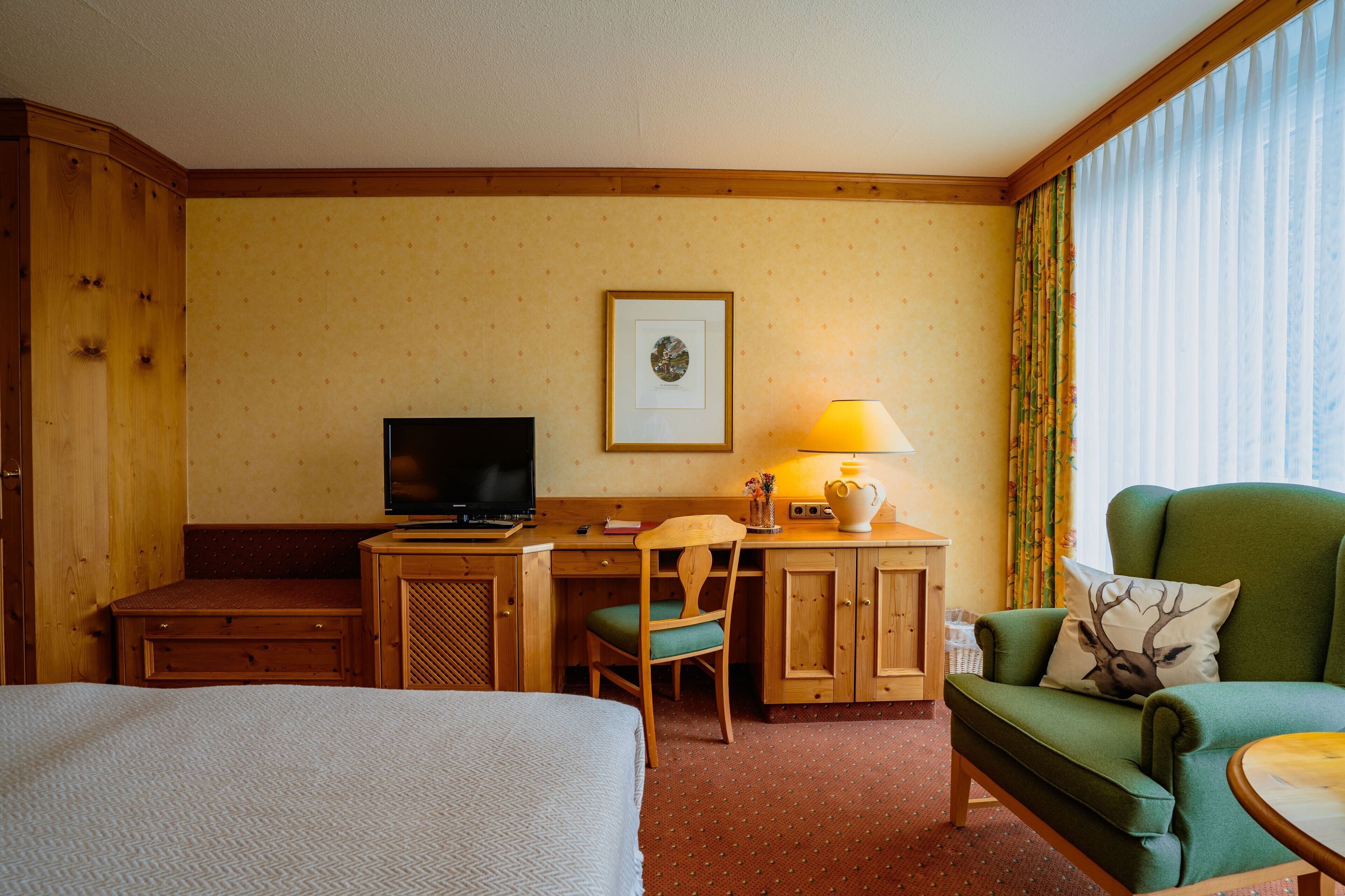 A hotel room with a bed, a desk with a television, a green armchair, and a window with curtains.