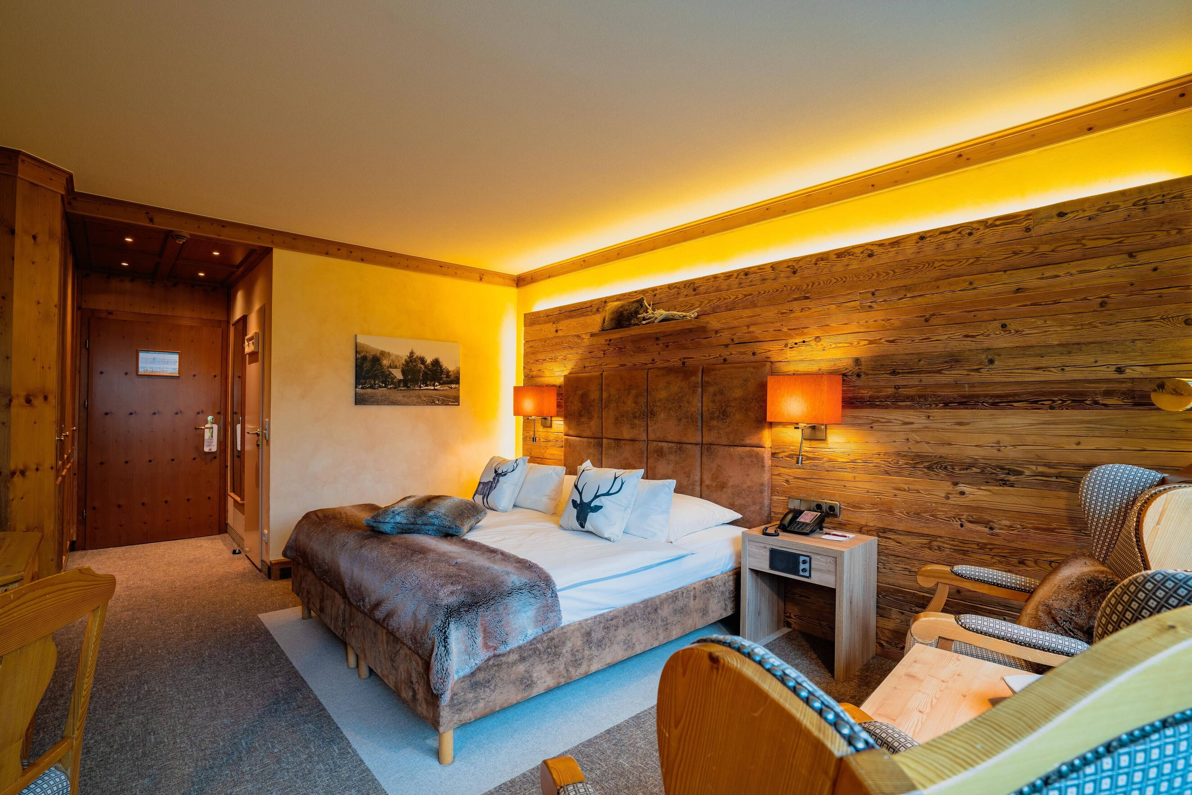 A bedroom with a wooden headboard, a bed with a white comforter, two bedside lamps, a nightstand, a chair, and a door.