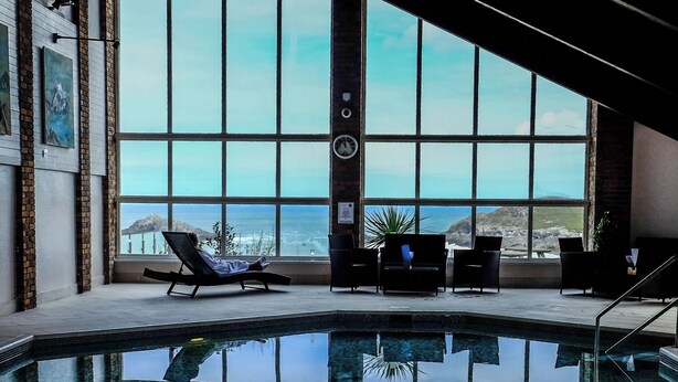Une piscine intérieure offrant une vue dégagée sur l’océan grâce à de grandes fenêtres, entourée de chaises longues et dotée d’un sol carrelé.