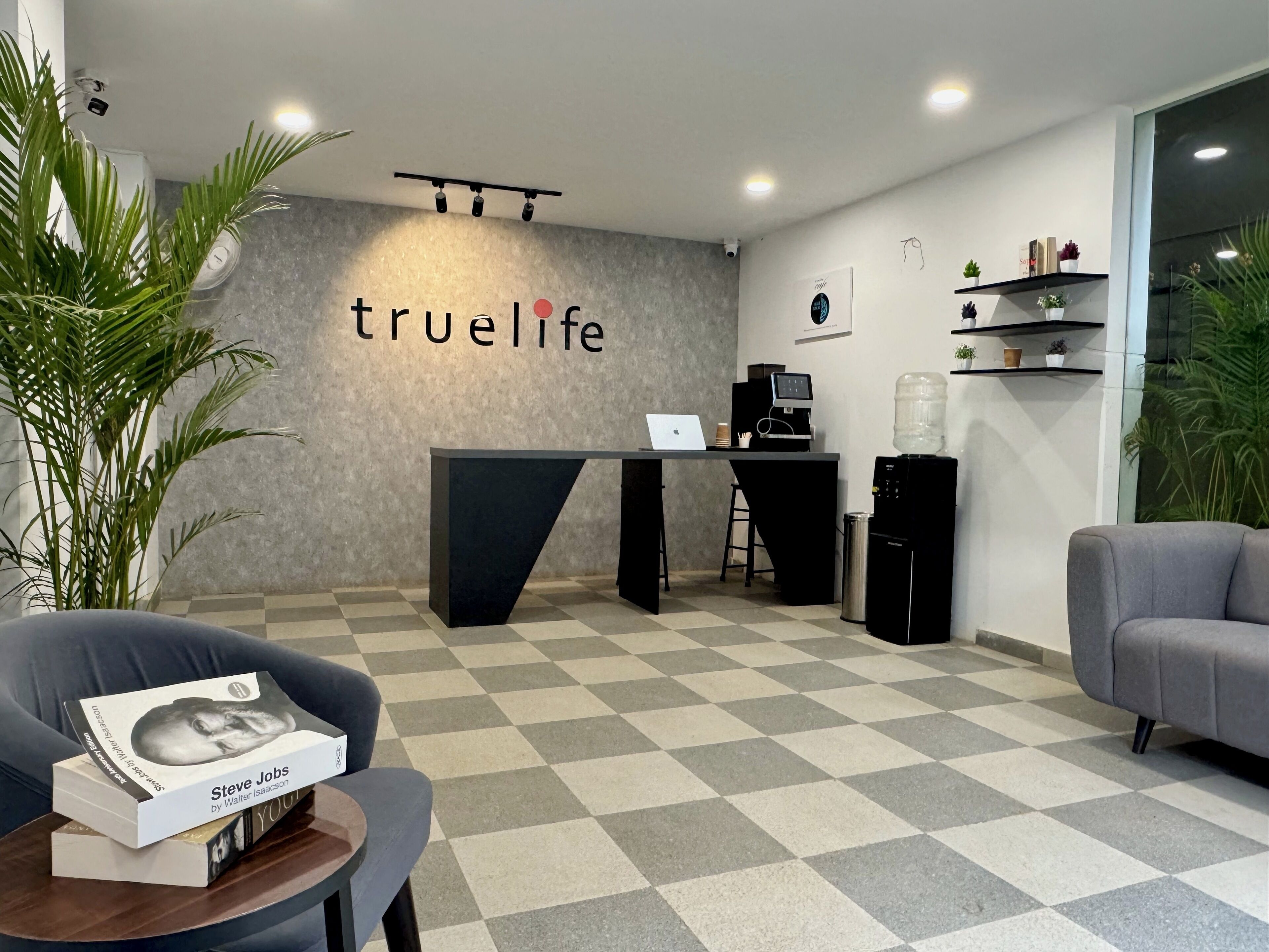 A reception area with a checkered floor, a reception desk, a computer, and a water dispenser. There are potted plants and a grey sofa.