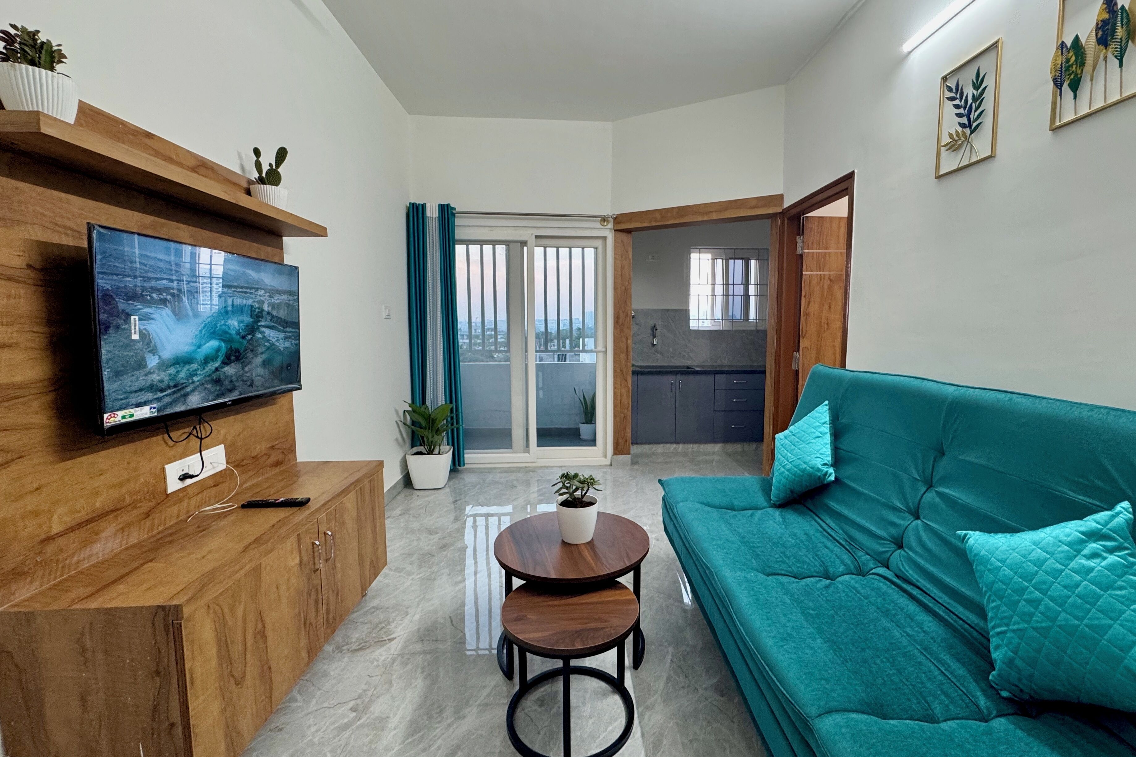 A modern living room with a flat-screen TV, wooden cabinet, teal sofa, and a small round table with a plant.