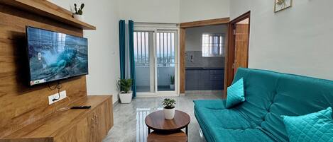 A modern living room with a flat-screen TV, wooden cabinet, teal sofa, and a small round table with a plant.