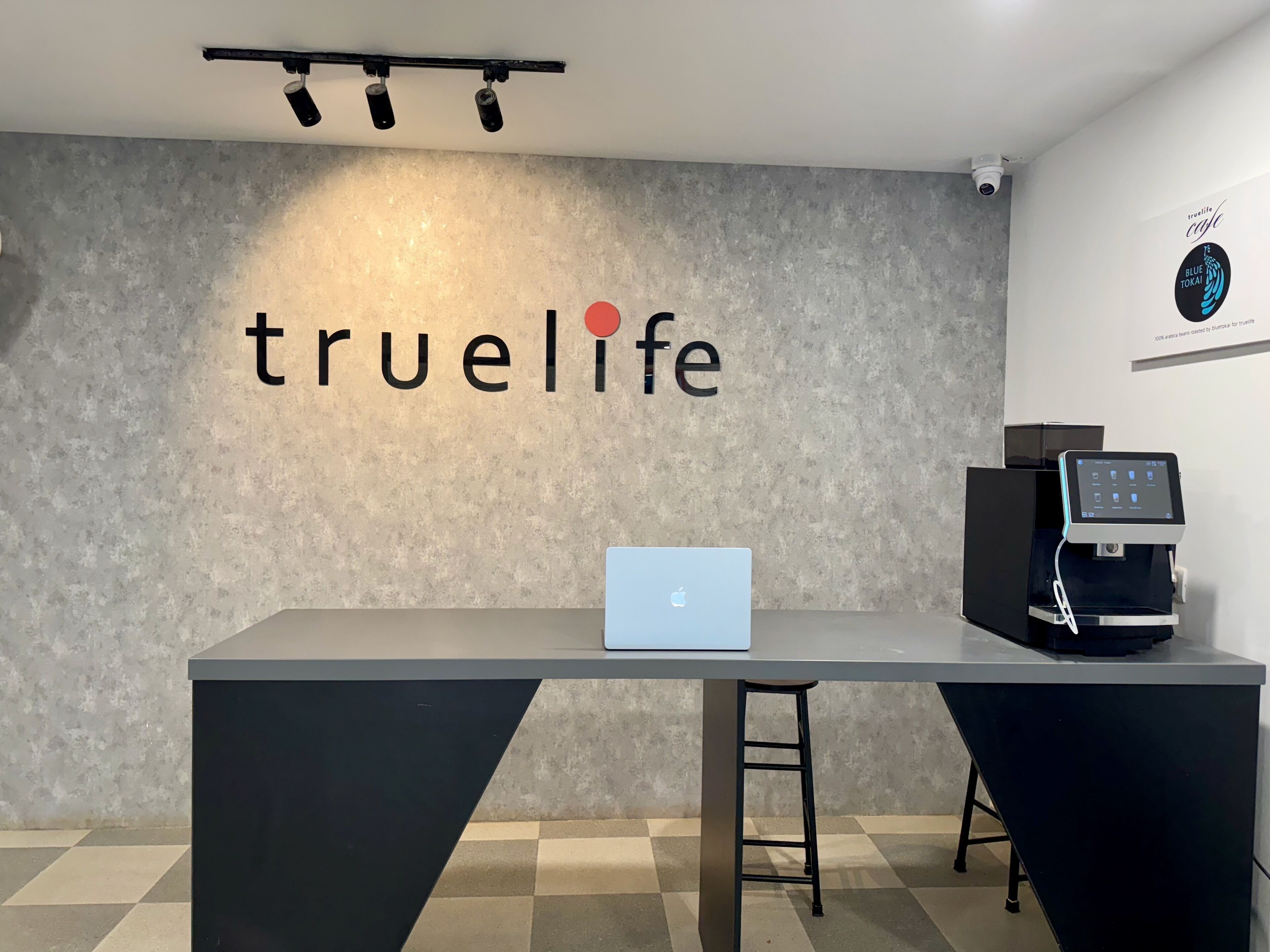 A reception desk with a laptop, a coffee machine, and a wall with the text "truelife".