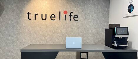 A reception desk with a laptop, a coffee machine, and a wall with the text "truelife".