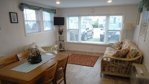 A dining area with a wooden table and chairs, a sofa, and a TV mounted on the wall. - Charming 1-bedroom cottage in beautiful Narragansett with AC, WiFi (Narragansett)