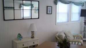 A room with a wooden table, a white dresser with wicker baskets, a wicker chair, a wall-mounted mirror, and a window with a curtain. - Charming 1-bedroom cottage in beautiful Narragansett with AC, WiFi (Narragansett)