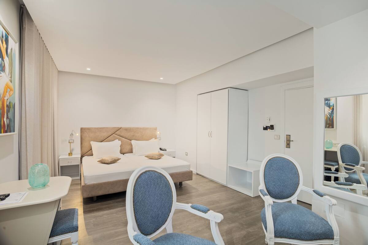 A modern hotel room with a bed, two chairs, a desk, and a wardrobe.