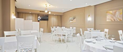 A spacious dining area with white tables and chairs, a flat-screen TV mounted on the wall, and a large abstract painting on the wall.