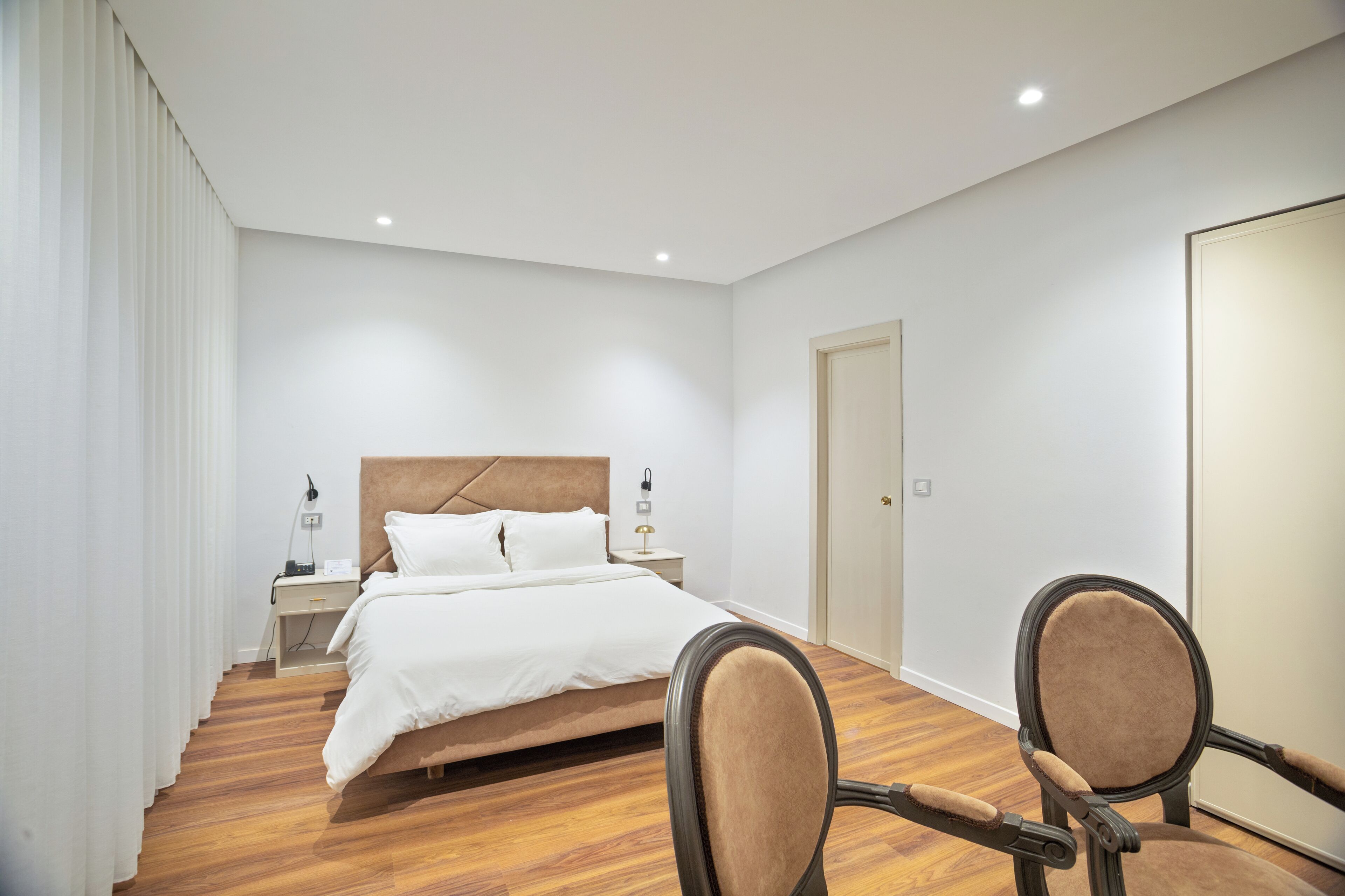 Una habitación de hotel moderna con una cama bien hecha, suelo de madera y dos sillones tapizados.