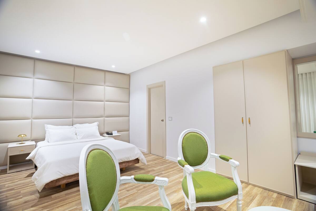 A modern bedroom with a large bed, two green upholstered chairs, a nightstand, and a wardrobe.