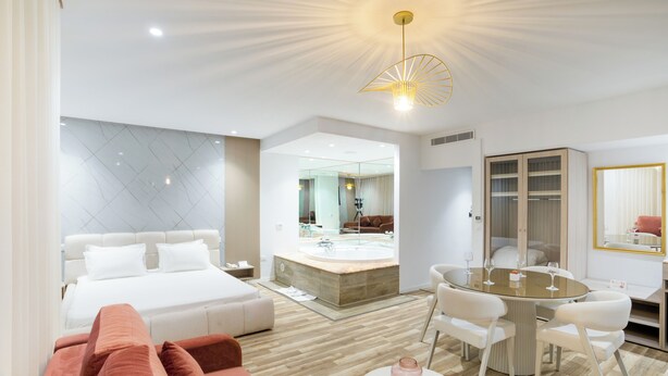 A modern hotel room with a bed, a sitting area with a sofa, a dining table with chairs, a large mirror, and a ceiling light fixture. - Hotel Donia Sfax (Sfax Ouest)