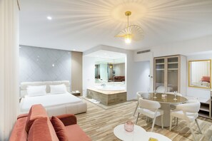 Una habitación de hotel moderna con cama, un área de descanso con sofá, una mesa de comedor con sillas, un espejo grande y un lámpara en el techo.