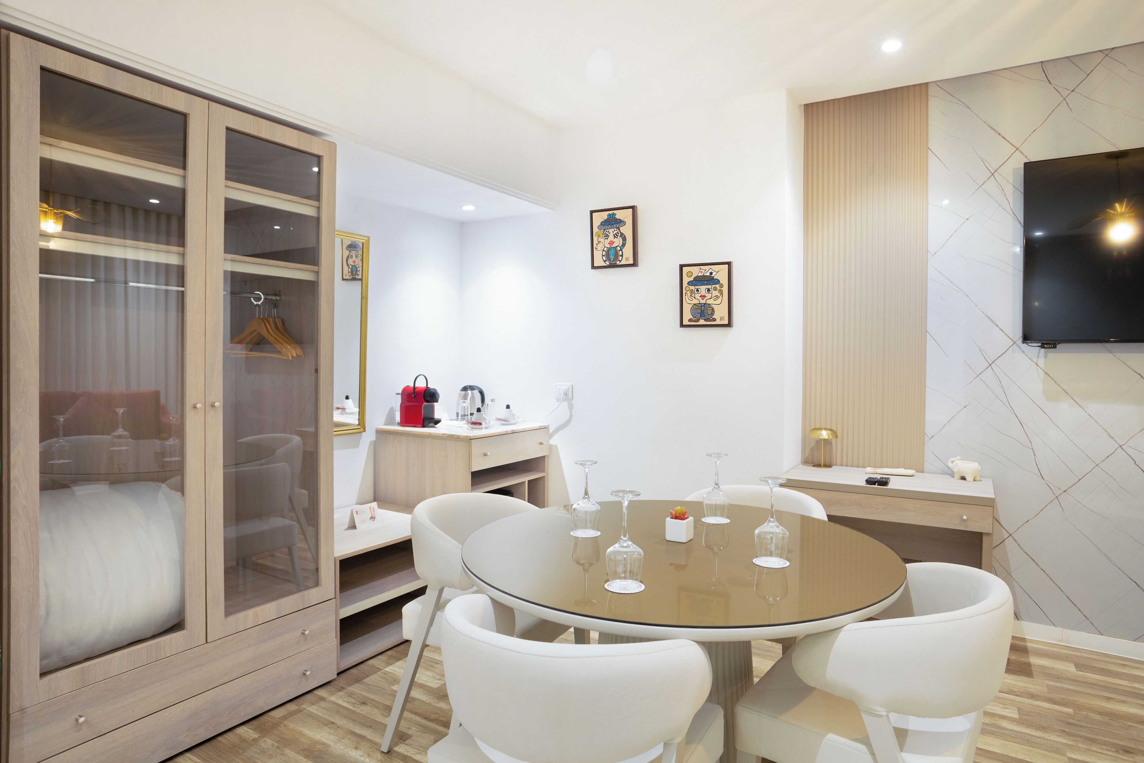 Una habitación moderna con una mesa redonda de vidrio, sillas blancas, un armario con puertas de cristal, un televisor de pantalla plana montado en la pared y dos cuadros enmarcados.