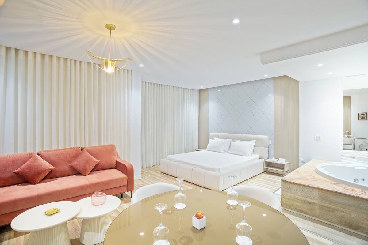 A modern hotel room with a large bed, a sitting area with a sofa and coffee table, a dining table with wine glasses, and a separate area with a hot tub.