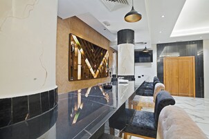 Un moderno vestíbulo de hotel con un mostrador de recepción alargado, un muro decorativo de madera, un pilar grande y una zona de estar con un sofá y reposapiés.