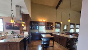 Una cocina amplia con encimeras de granito, armarios de madera y una isla central. Se observan luces colgantes, un refrigerador de acero inoxidable y un amplio ventanal que proporciona luz natural.