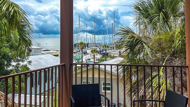 Un balcón con dos sillas de mimbre y una mesa, con vistas a un puerto deportivo con barcos y palmeras.