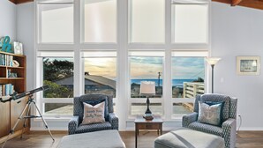 Ein Zimmer mit zwei gemusterten Sesseln, einem Holzregal, einem Teleskop und Meerblick durch große Fenster.