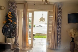 Une porte ouverte donnant sur un patio avec vue sur la mer, un ventilateur, un pot de fleurs contenant des tournesols et un vase avec des fleurs blanches.