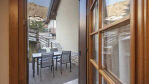 A view through a glass door of a dining area with a table and chairs, a stone wall, and a staircase leading to a building with a sloped roof. - Silia's Home - Two Bedroom Apartment, Sleeps 4 (Vione)