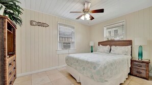 Un dormitorio con cama, dos mesitas de noche, ventilador de techo, ventana y un cartel en la pared.