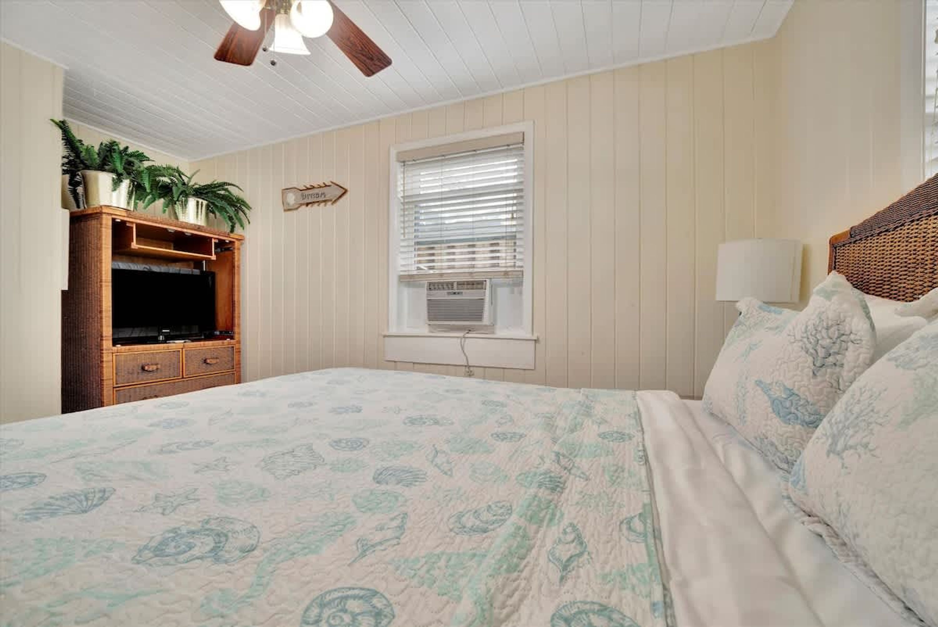 A bedroom with a bed, a wooden dresser, a ceiling fan, a window with blinds, and a wall-mounted air conditioner.