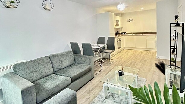 A modern living room with a grey sofa, a glass coffee table, and a small side table. There is a kitchen area in the background with white cabinets and a black countertop. - 2br-sleeps4-citycentre-parking-rooftop (Birmingham)