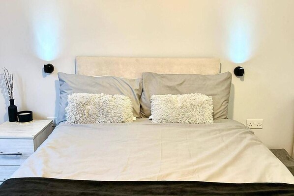 A bed with a white headboard and two fluffy pillows, a bedside table with a black vase and a small black object, and a wall-mounted light fixture. - 2br-sleeps4-citycentre-parking-rooftop (Birmingham)
