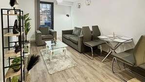 A modern living room with a grey sofa set, a glass coffee table, and a metal side table. There is a bookshelf with decorative items and a potted plant. - 2br-sleeps4-citycentre-parking-rooftop (Birmingham)