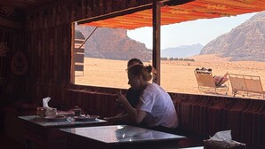 A room with a large window overlooking a desert landscape, a table with a tissue box, and a chair. - Wadi Rum Magic Nature Camp (Wadi Rum)
