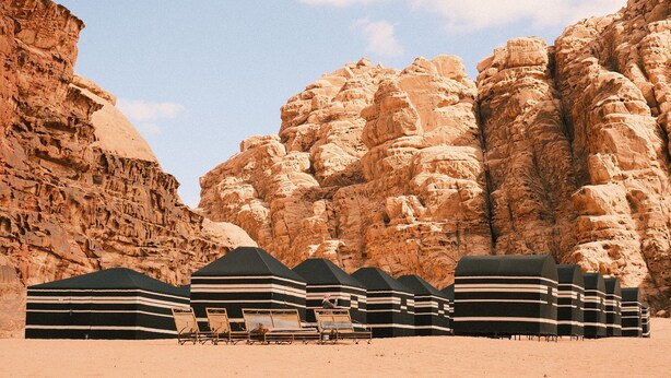 A desert landscape with rock formations and a row of striped tents with lounge chairs. - Wadi Rum Magic Nature Camp (Wadi Rum)