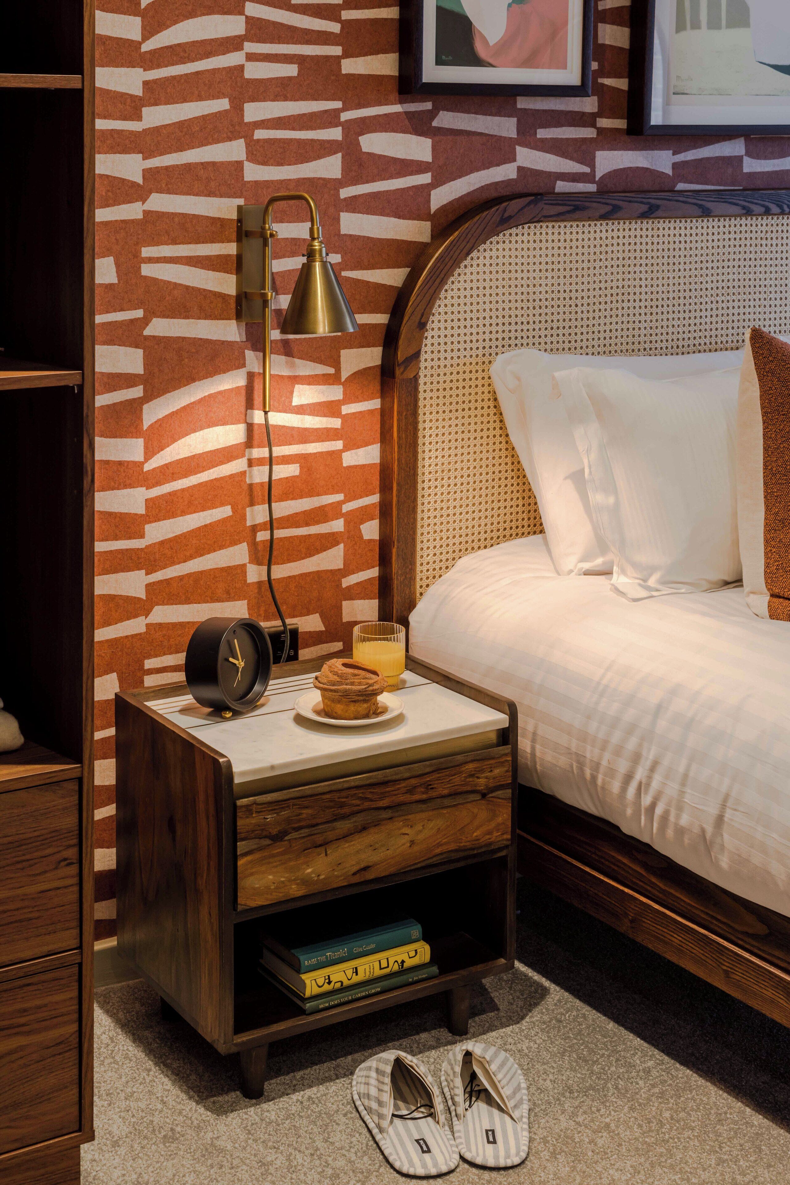 A neatly arranged bedroom with a wooden nightstand, a bedside table with a lamp, a bed with white bedding, and a wall with a patterned design.