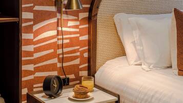 A neatly arranged bedroom with a wooden nightstand, a bedside table with a lamp, a bed with white bedding, and a wall with a patterned design.