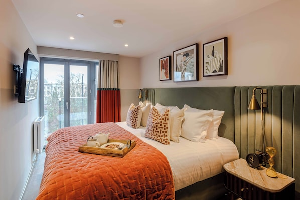 Una habitación de hotel con una cama grande, una mesita de noche con un reloj y una lámpara, un televisor en la pared y una ventana con cortinas.