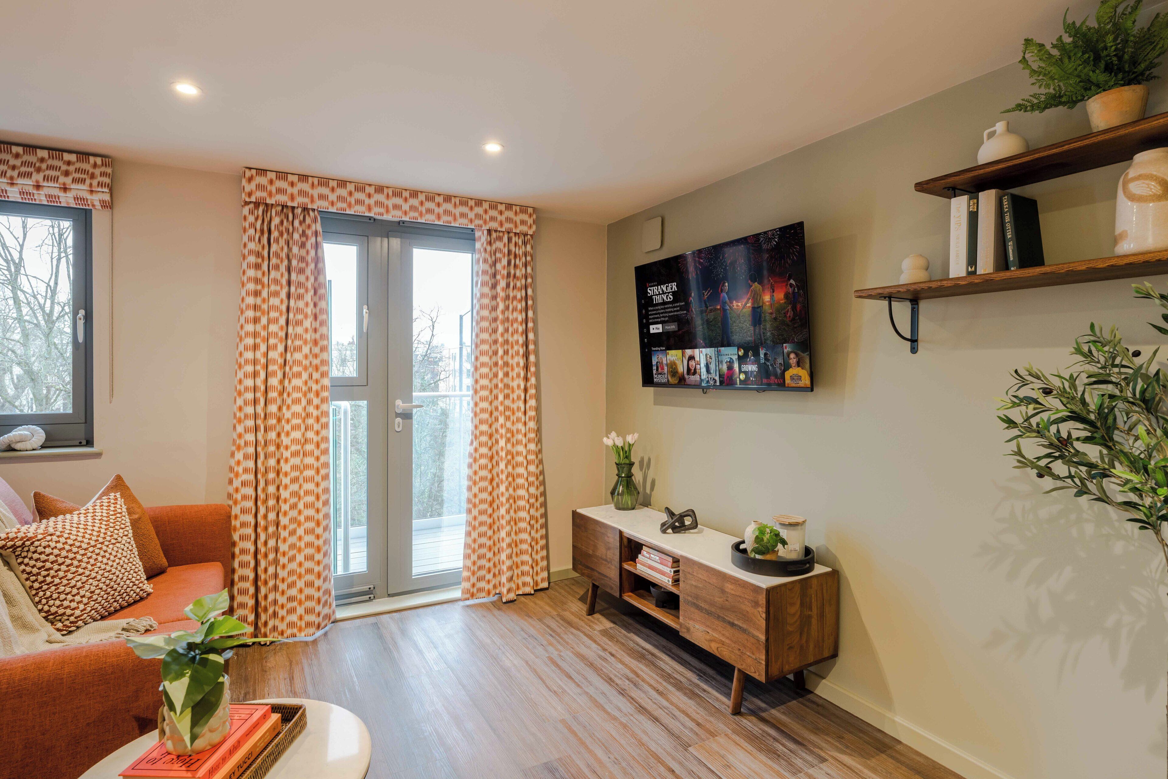 A living room with a sofa, a coffee table, a TV mounted on the wall, a wooden cabinet, and a window with curtains.