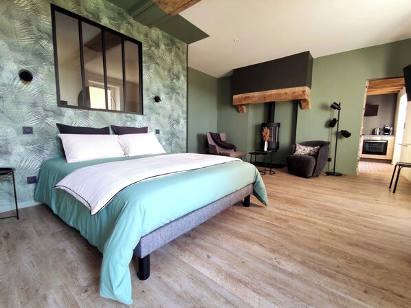 A bedroom with a bed, a chair, a small table, a wall-mounted mirror, and a built-in wood stove.