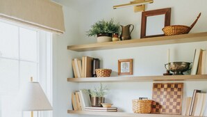 Una sala de estar acogedora con un sillón beige, una mesita redonda y una lámpara de pie. Estanterías sobre el sillón exhiben libros, un tablero de ajedrez y objetos decorativos.