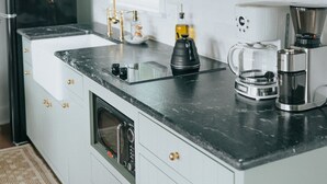 Una cocina moderna con encimera negra, armarios blancos, horno integrado, cafetera y lámpara colgante.