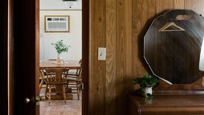 A wooden door leading to a room with a dining table, chairs, a potted plant, and a mirror on the wall.