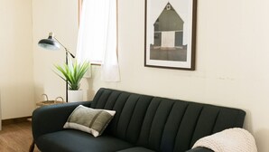 A dark green sofa with a single cushion, a floor lamp, a potted plant, and a framed picture of a barn on the wall.