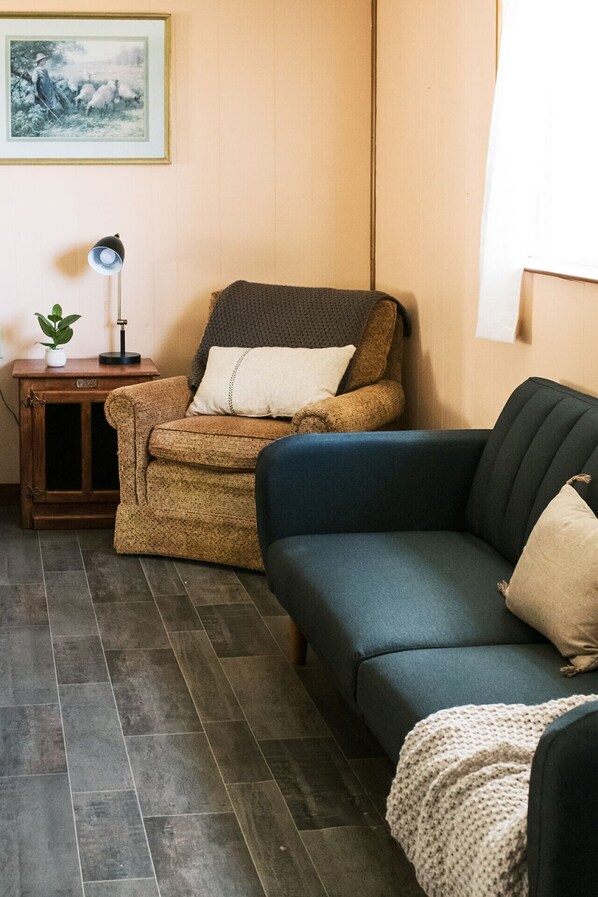 A cozy living room with a brown armchair, a blue sofa, a small wooden side table with a lamp, and a framed picture on the wall.