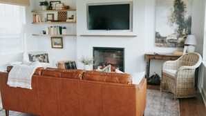 Un salon avec un canapé en cuir marron, un téléviseur à écran plat, une cheminée, des fauteuils en osier et des étagères remplies de livres et d’objets décoratifs.